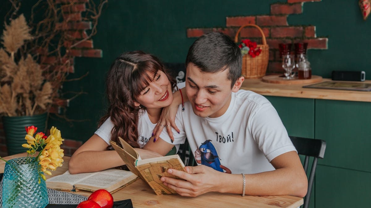 Positive Asian girlfriend hugging boyfriend while reading book together sitting at table in kitchen